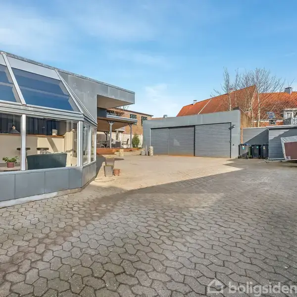 En gårdsplads med flisebelagt grund, hvor en stor glaskonstruktion, muligvis en udestue, ses til venstre. Til højre står en metaltrailer foran en grå garagebygning med rulleporte.