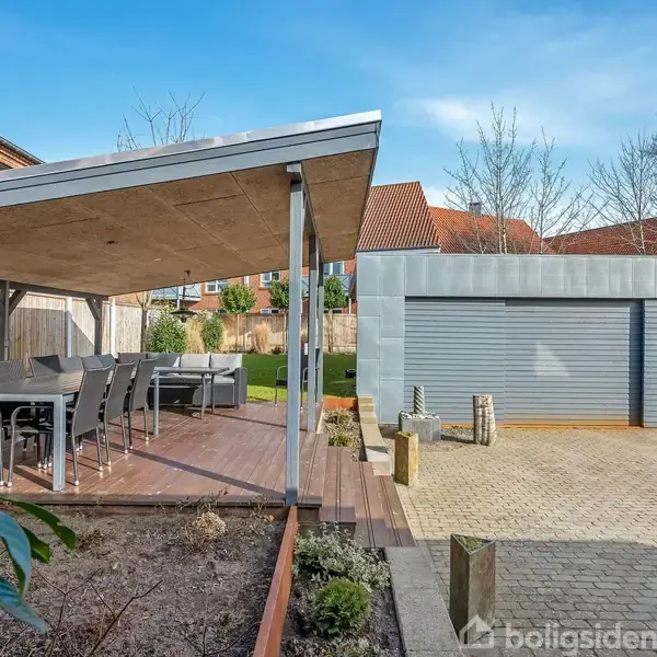 Et overdækket terrasseområde med spisebordssæt, omringet af hegn og planter. Ved siden af står en bygning med to grå garageporte, omgivet af brostensbelægning og haveområde.