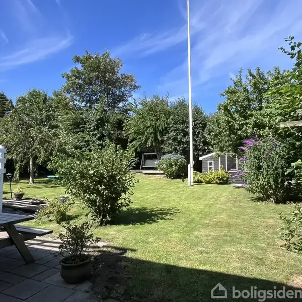 Græsplæne med træer og buske omkranset af have, set fra terrasse med bænk. I baggrunden står en flagstang og lille haveskur. Himlen er blå med skyer.