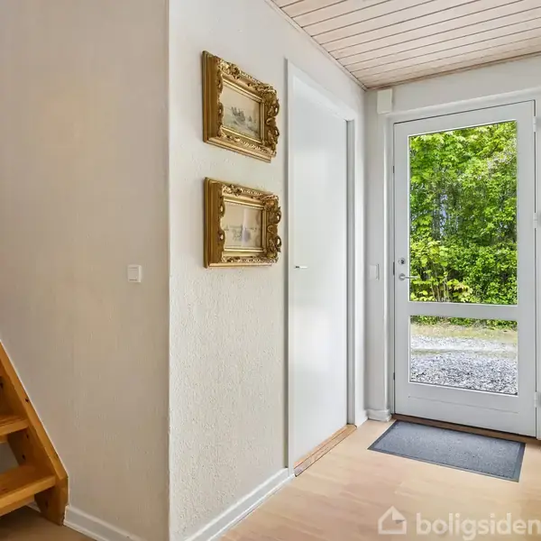 Trætrappe står ved siden af en hvid væg med to indrammede billeder, mens en glasdør åbner til en have med frodig bevoksning udenfor.