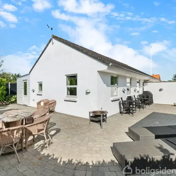 Et hvidt hus står i en solrig have med en terrasse belagt med fliser. Havemøbler og borde er opstillet, flankeret af buske og træer under en blå himmel.