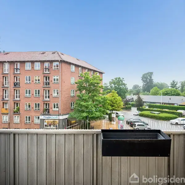 Balkon med blomsterkasse, beliggende foran et boligkompleks. Det regner, og mennesker med paraplyer bevæger sig på et hjørne af en stille gade omgivet af træer og biler.