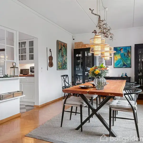 Et træbord med stole omkring står i et spisestueområde. Bordet er pyntet med en vase med blomster, omgivet af kunstværker og glasvitrineskabe. Rummet er lyst med trægulv.