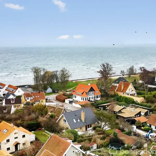 Flere huse med røde tage står i en landsby nær en kystlinje. Havet ligger i baggrunden med få skyer på himlen, og træer grænser området.