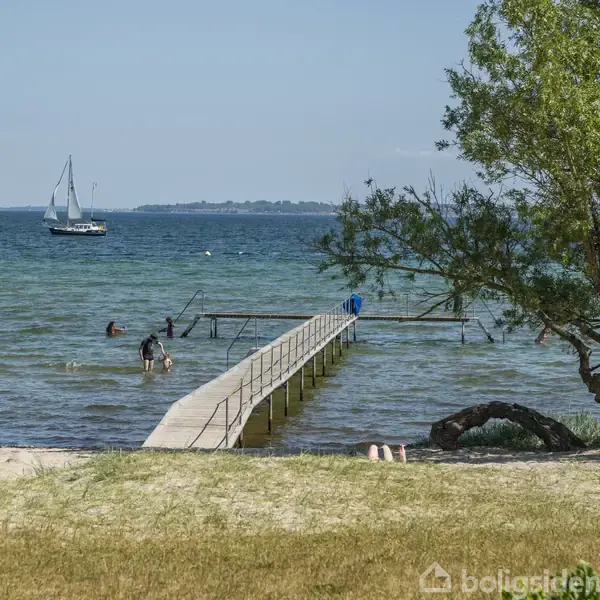 En træbro strækker sig ud i en klar blå sø, hvor folk bader, mens en sejlbåd ses i det fjerne. Træer og strandgræs omkranser området.