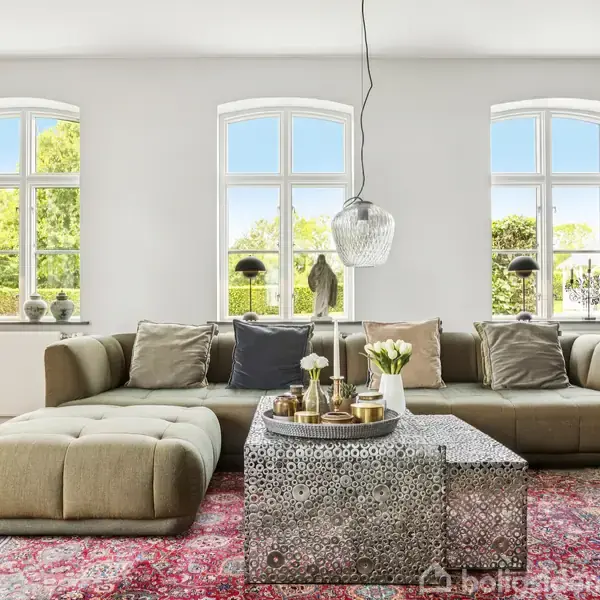 Store, grønne sofaer står ved et dekorativt bord med blomster og lys, placeret på et orientalsk tæppe i en lys stue med tre buede vinduer og udsigt til træer.