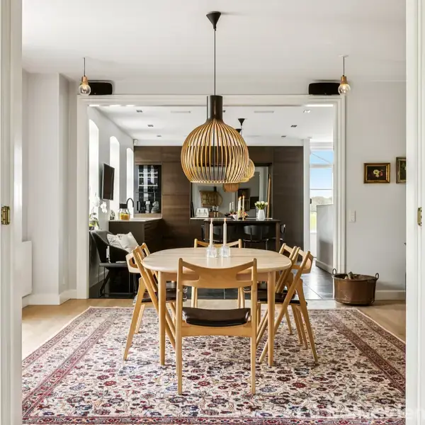 Rundt spisebord med fire stole står under en stor, hængende lampe. Rummet er lyst og moderne med åben passage til køkkenet i baggrunden. En tæppe dækker gulvet.