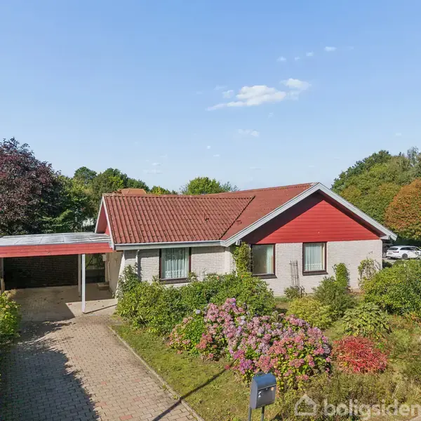 En étplanshus med rødt tag og træfacade ligger omgivet af en velplejet have med buske og blomster. En carport ligger til venstre, omgivet af grønne træer og buskadser.