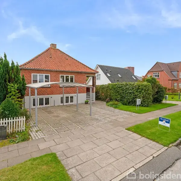 Rød murstenshus står tomt med en dobbelt carport foran. Huset er placeret i en forstad med græsplæner og hække langs vejen. Skilt: "Til salg - EDC Poul Erik Bech".