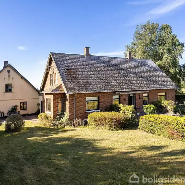 Et rødt murstenshus med skråtag står i en velplejet have omgivet af buske. Foran huset er en græsplæne, og i baggrunden ses et mindre, lignende hus.