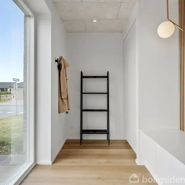 Sort reol står i en lys entre med trægulv, ved siden af en frakke på en krog. Stor rude viser vej med græsplæne udenfor, og en moderne lampe hænger i loftet.