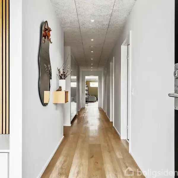 Gang med trægulv strækker sig i et moderne hus. En kunstnerisk spejl og hylde pryder venstre væg, mens flere døre ses langs højre væg. Loftet har indbyggede lys.