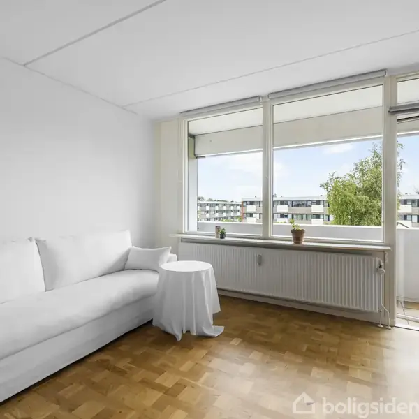 En hvid sofa står i et lyst stuehjørne. Et lille bord ved siden af sofaen. Store vinduer og en åben altandør giver udsigt til træer og bygninger.