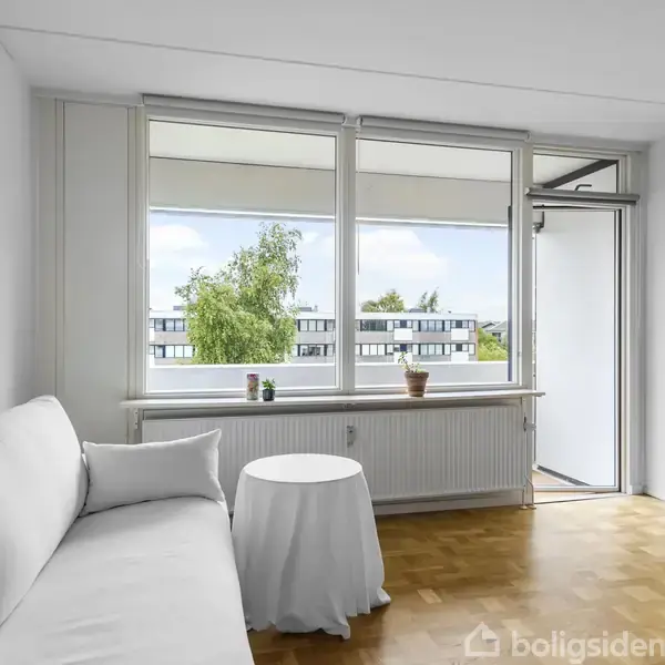 En hvid sofa står ved siden af et rundt bord med dug, placeret i en lys stue med trægulv. Store vinduer viser ud til en balkon og et træ udenfor.