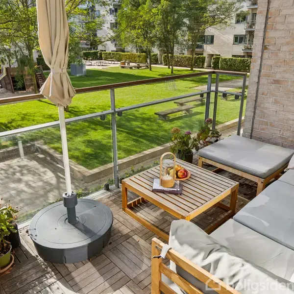 Terrassemøbler står på en altan med grå hynder og en parasol. Der er et lille bord med frugt og kande. Udsigt til en grøn plæne med træer og lejlighedsblokke.