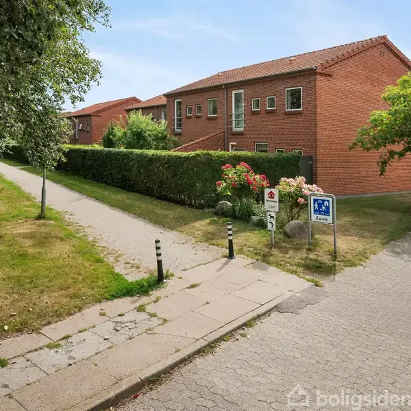 Rød murstensbygning ses langs en asfalteret vej. En sti og buskads flankerer bygningen. Skilt med teksten "25 Sone" og et andet skilt er synlige i forgrunden.