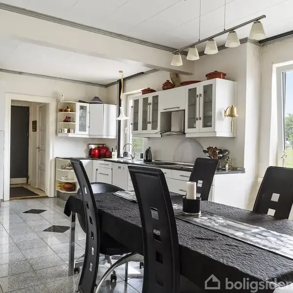 Spisebord står i et lyst køkken med sorte stole omkring. Køkkenskabe og apparater fylder baggrunden, mens et vindue viser en udsigt til grønt landskab udenfor.
