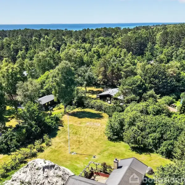 Grønt område med huse omringet af træer. Hovedhus i nederste del, græsplæne i midten. Skov i baggrunden, og hav i horisonten. Ingen synlig tekst.