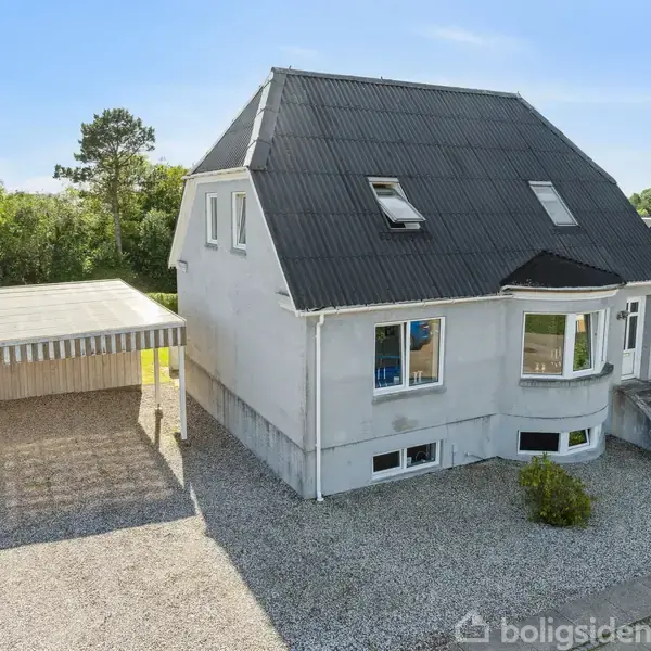 Et gråmalet hus med sort tag står i en grusbelagt indkørsel ved siden af en carport. Trapper fører til hoveddøren. Omgivet af grønne buske og træer.