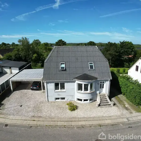 Et hvidt hus med gråt tag står på en parcel med grusbelagt forhave. En carport med bil ved siden af. Omgivet af grønt område og naboer på en stille vej.