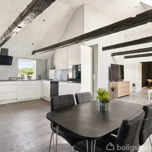 En moderne stue med synlige bjælker og skrå lofter. Et sort spisebord med stole står i midten, mens et lyst køkken ses bagved. En stue med sofaer er i baggrunden.