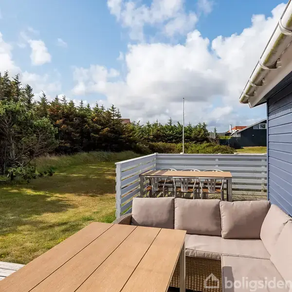 Træbord med beige sofa står på terrasse ved blåmalet hus. Omkring er grønt græs og tæt bevoksning af træer under en delvis skyet himmel.