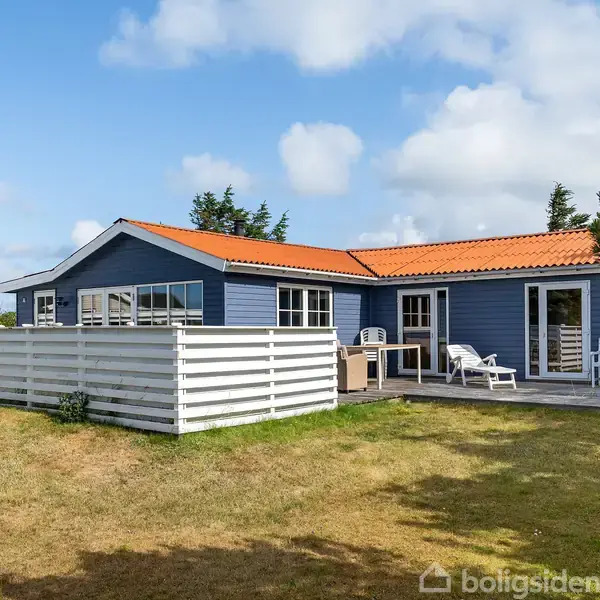 Et blåt sommerhus med rødt tag står i en have med græsplæne. Foran er der en terrasse med havemøbler. Flere træer kan ses i baggrunden under en blå himmel.