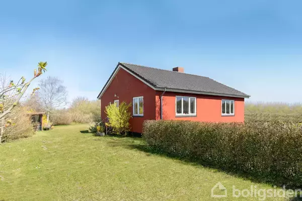 Rødt hus står stille på en grøn græsplæne omgivet af buske og træer under en klar blå himmel.