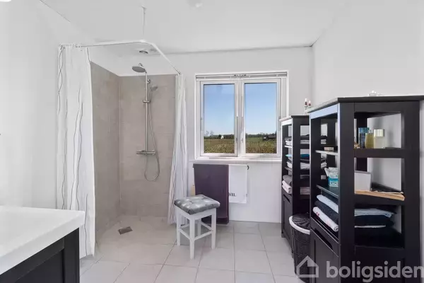 Badeværelse med bruser til venstre, en stol i midten og to åbne hylder fyldt med håndklæder og toiletartikler til højre. Vindue viser landskab udenfor.