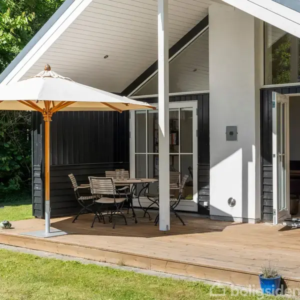 Terrasse med parasol og træmøbler placeret foran et moderne sommerhus med store vinduer. Omgivet af frodig grøn have baggrund.