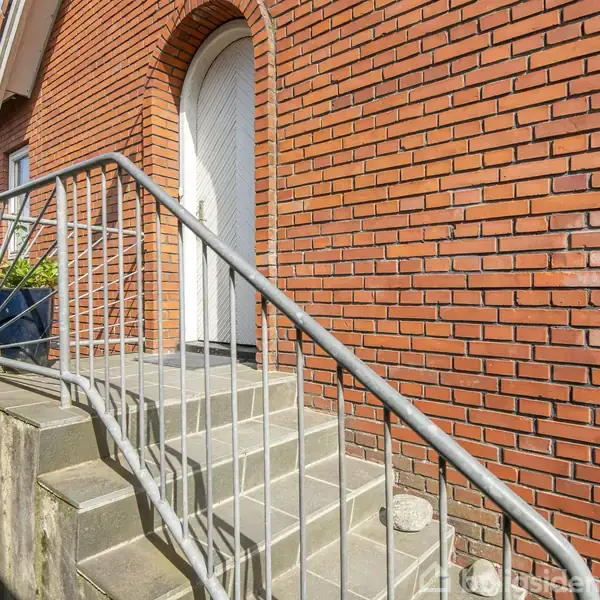 Røde murstensbygning med en hvid dør, flankeret af en trappe med metalgelænder. Trappen fører til en forhøjet indgang. Omkringliggende miljø viser græsset område og flisebelagt sti.