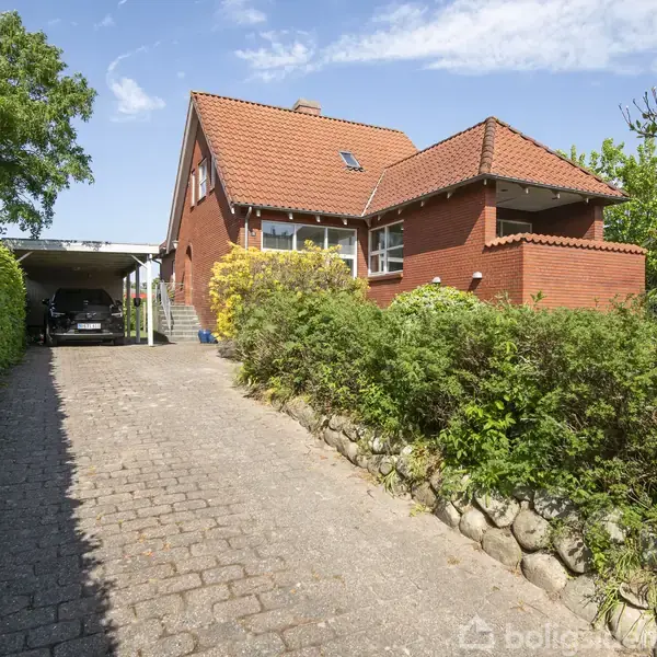Rødt murstenshus med rødt tag står i en grøn have. En brolagt indkørsel fører op til carporten og huset. Solskin og blå himmel ses i baggrunden.