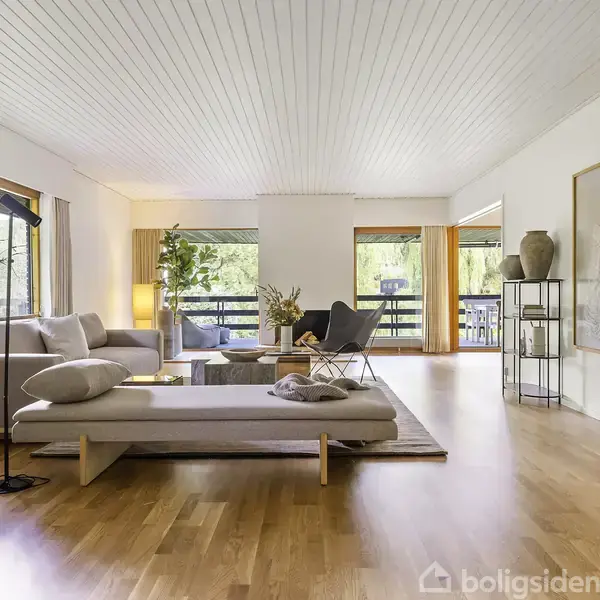 Stue med moderne sofa og chaiselong, placeret på lyst trægulv. Store vinduer omgiver rummet, der er dekoreret med planter og kunstværker, og tilbyder udsigt til en balkon og grønne omgivelser.