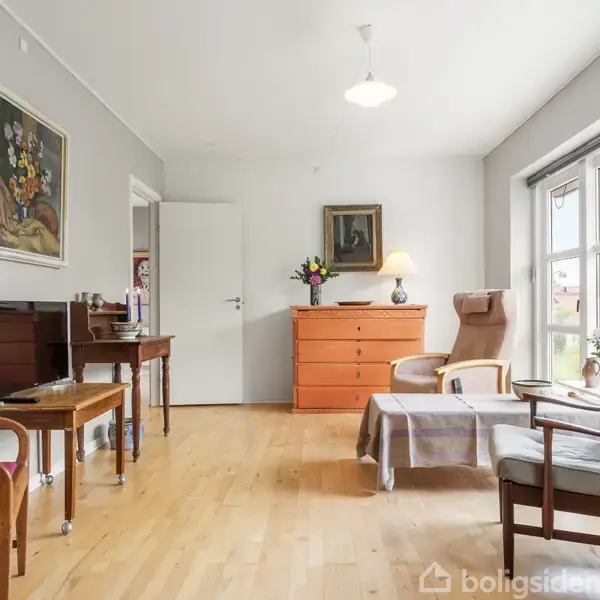 Stue med træmøbler, hvor en orange kommode står op ad en hvid væg, flankeret af lænestole og et bord. Store vinduer slipper lys ind, og vægkunst pryder rummet.