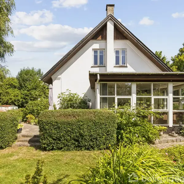 Et hvidt hus har en glasveranda foran og ligger i en have med grønne buske og træer. Himlen er blå med lette skyer.