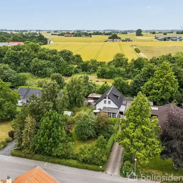 Hus omgivet af frodige træer og have ses fra oven, placeret i landligt område med marker og spredte bygninger i baggrunden under en blå himmel.
