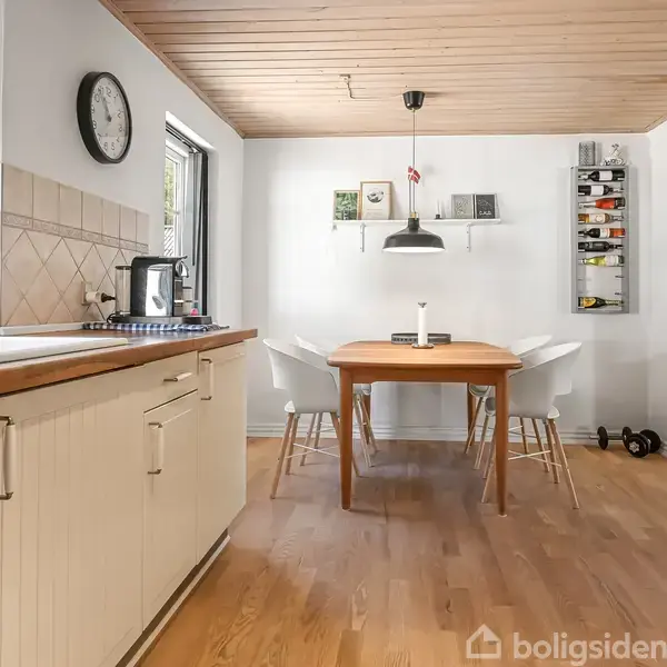 Køkken med træbord omgivet af fire stole. Bordet står under en hængende lampe. Til venstre er en hvid køkkenvask ved siden af et vindue. Væg med hylder og pynt.