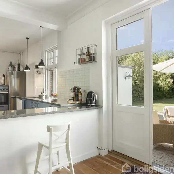 Køkken med hvid stol ved barbord, åbner til terrasse med udendørs møbler og parasol i haveomgivelser. Lyse træ- og hvide designelementer skaber en åben og indbydende atmosfære.