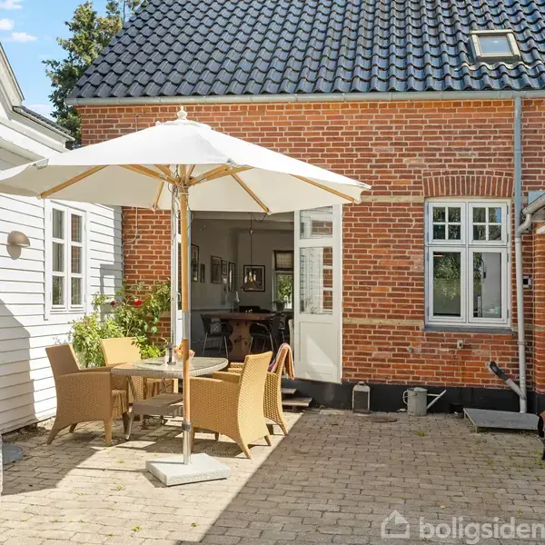 En gårdsplads med havemøbler og en parasol, hvor et rødt murstenshus og et hvidmalet træhus omkranser området, skaber en hyggelig udendørs atmosfære under en solrig himmel.