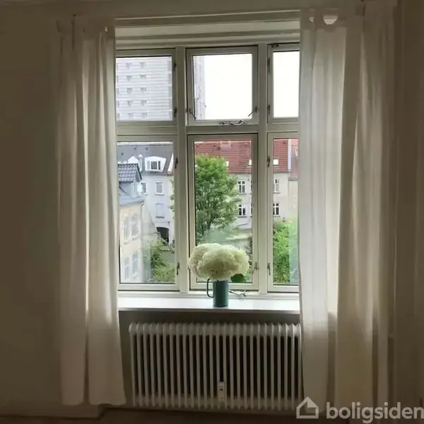 En vindueskarm med en vase med hvide blomster. Vinduet åbner mod en udsigt over bygninger og træer. Gardiner hænger på begge sider, delvist tilbagetrukket.