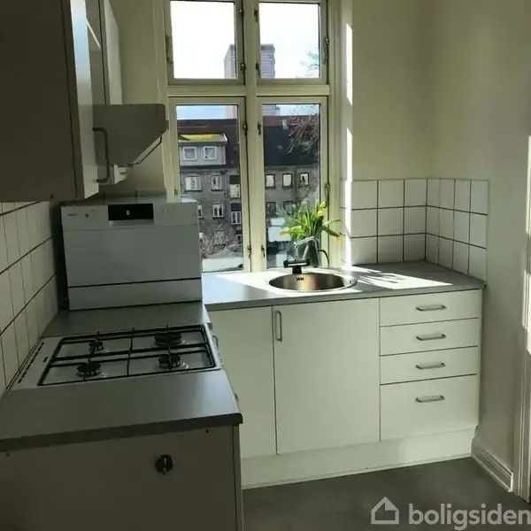 Et køkken med en gaskomfur og ovn, står i et lille rum ved et vindue med udsigt til bygninger. En plante står ved vasken på en lys bordplade.