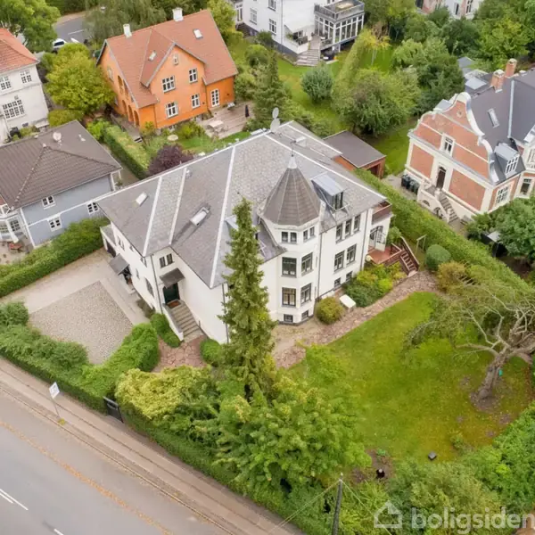 Stort hvidt hus med spidst tårn står stille på en velplejet grøn grund, omgivet af andre villaer og træer langs en vej.