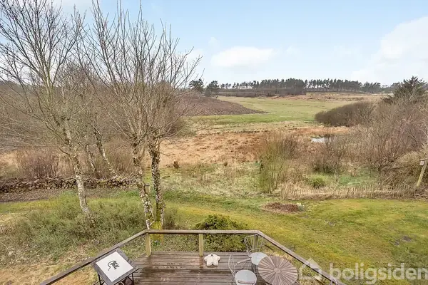 Træterrasse med bord og stole står stille i en have, omgivet af græs og træer, med udsigt over åbent landskab og en skov i baggrunden.