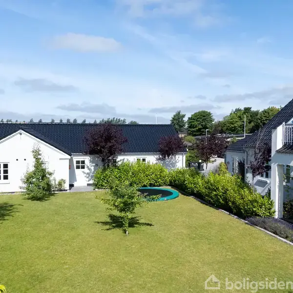 Et stort, hvidt hus med sorte tagsten står i en velplejet have med grøn græsplæne og en nedgravet trampolin. Træer og buske omgiver boligen under en blå himmel.