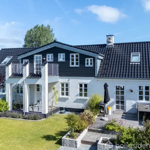 Et stort, hvidt hus med sort tag står i en velplejet have. Huset har flere altaner og høje vinduer. Himlen er klar og blå, og der er en terrasse med planter.