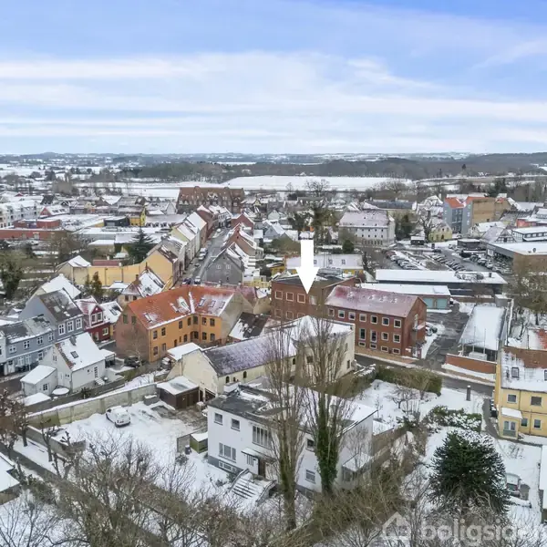 By i sneklædt landskab med mange tætliggende huse og røde tage. Hvid pil viser mod et specifikt hus. Sne dækker tagene og jorden, omgivet af træer og bygninger.