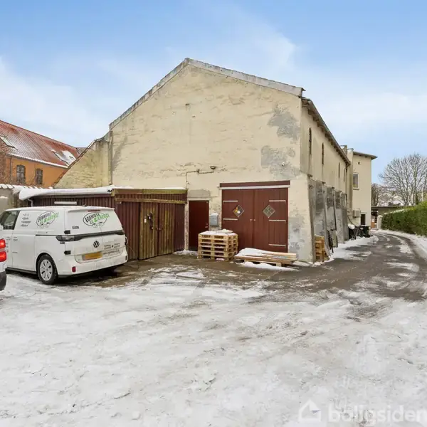 En hvid varevogn holder på en snebelagt gårdsplads foran en ældre, gulmalet bygning. Bygningen har en parkeret palle foran og er omgivet af lave hegn og træer. Tekst på varevognen: "Egemose VVS & Energi".