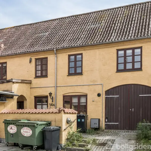 Gul murstensbygning med to etager og brune døre og vinduer, hvoraf én dør bærer nummeret 14A. Foran bygningen står en grøn affaldscontainer i en brostensbelagt gård.