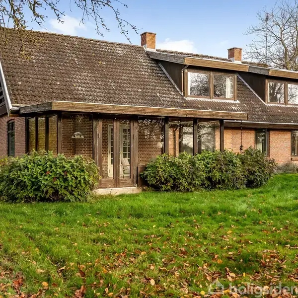 Et stort etplanshus med teglstensvægge og skråt tag står i en have. Huset har store vinduer og buske foran. Græsplænen omgiver huset, og der er spredte træer i baggrunden.