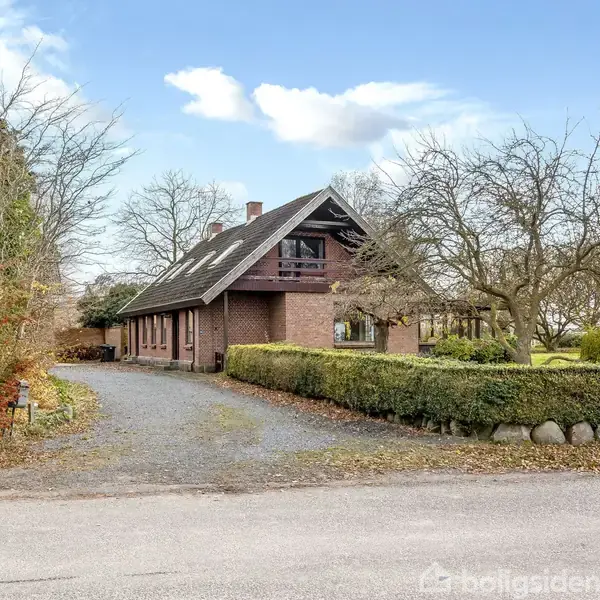 Et stort murstenshus med gavltag står på en grusvej, omgivet af en velholdt have med træer og buske. Et hegn og græsplæne omgiver huset.